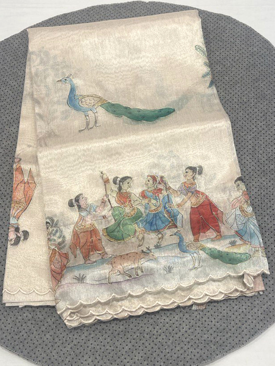A cream-colored linen tissue saree with a metallic gold sheen, featuring hand-painted Kalamkari motifs of traditional Indian dancers and peacocks along a scalloped border.