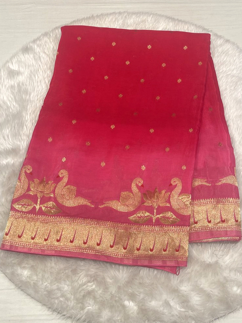 Fuchsia pink Malai silk saree featuring gold Zari butti work and a broad border with intricate woven swan and lotus motifs, displayed on a white faux fur rug.