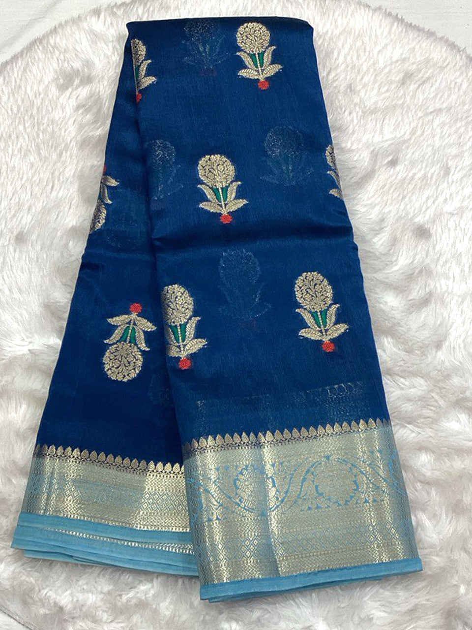 Royal blue semi-silk saree with gold mango floral motifs and a contrasting sky blue woven Zari border on a white faux fur background.