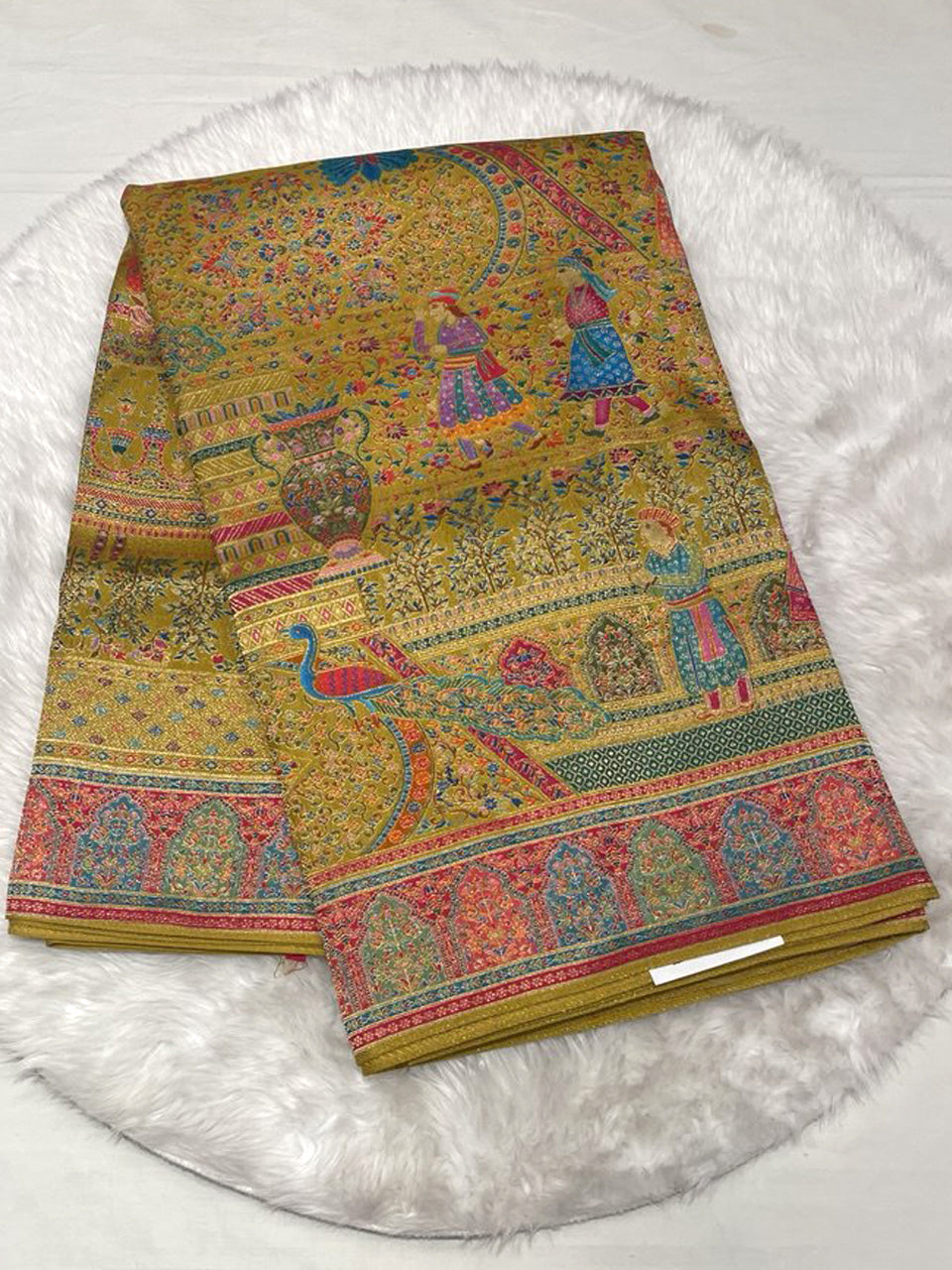 A folded handloom Banarasi Jamdani silk saree. The fabric is a mustard/olive green with intricate multi-color woven motifs depicting Rajasthani figures, peacocks, and vases. It features a wide, ornate pink and blue woven border.