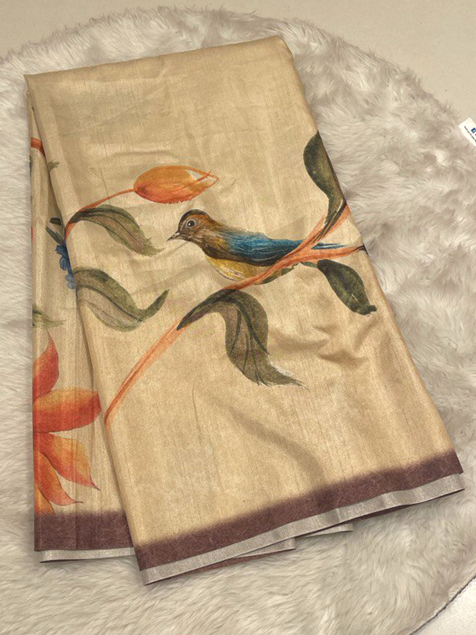 A light beige raw silk saree showcasing a nature-inspired print of a small bird perched on a branch with orange floral buds, featuring a dark chocolate brown contrast border and a subtle metallic sheen.
