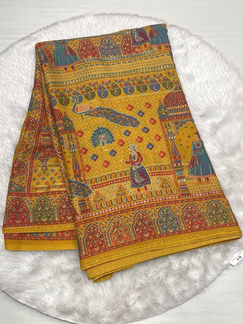 A folded mustard/golden yellow crepe saree featuring an all-over print of royal figures, peacocks, and architectural elements inspired by Banarasi designs. The wide border is multi-colored and heavily patterned.