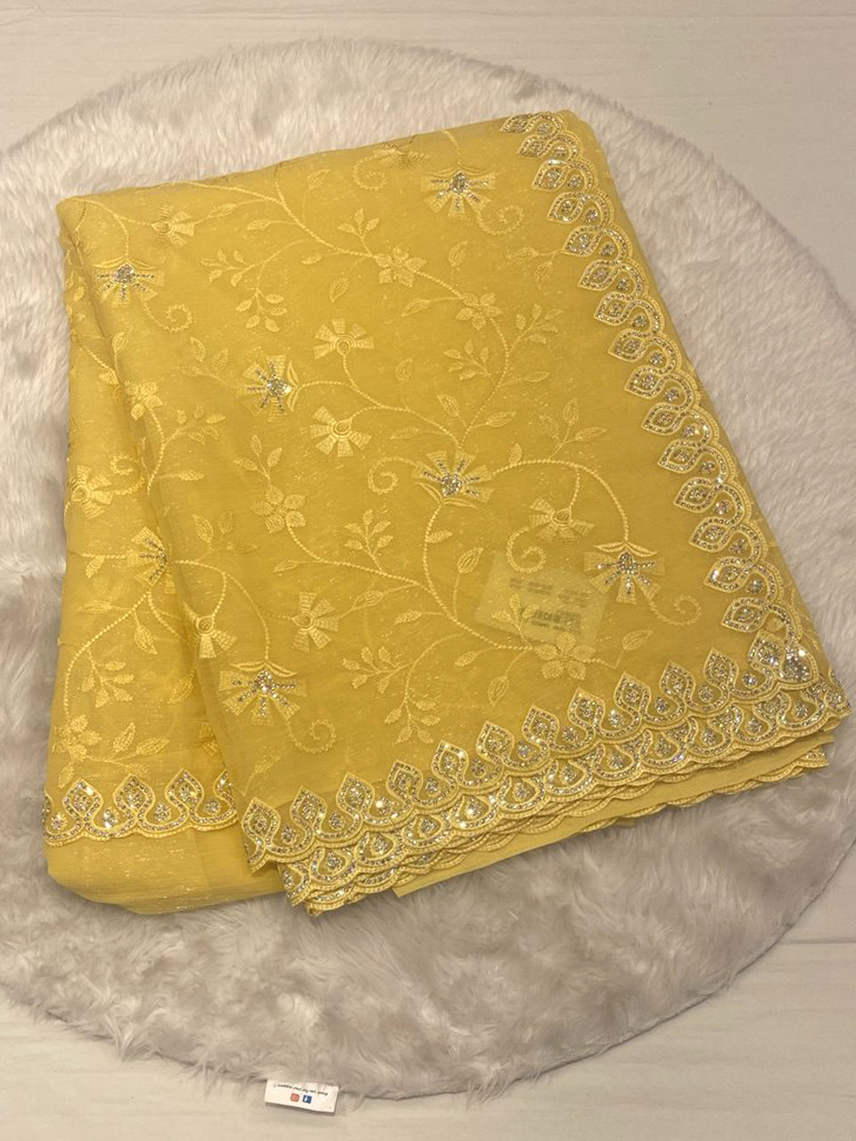 Folded lemon yellow shimmer georgette saree featuring tonal floral embroidery and a wide scalloped border embellished with silver sequins and stones, displayed on a white faux fur rug.