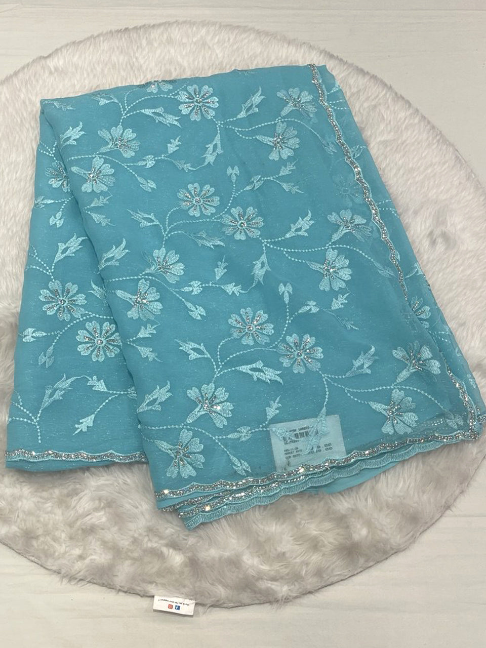 A sky blue shimmer georgette saree folded on a white faux fur rug. The saree features intricate tone-on-tone floral embroidery with delicate silver sequin accents throughout the body. It includes a decorative silver scalloped cut-work border with fine threadwork and stone embellishments